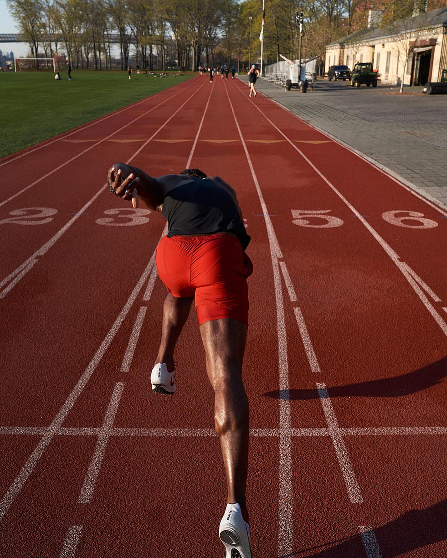 A sprinter launches from the starting blocks on a red running track, showcasing power and athletic form against a backdrop of green fields and trees.