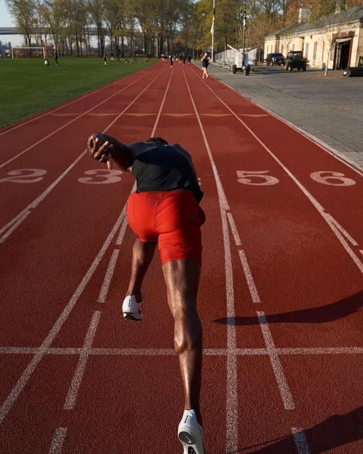 A sprinter launches from the starting blocks on a red running track, showcasing power and athletic form against a backdrop of green fields and trees.