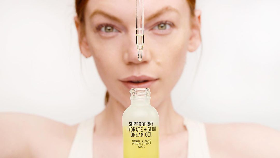 Close-up studio shot of a woman with red hair and pale skin presenting a bottle of Superberry Hydrate + Glow Dream Oil.