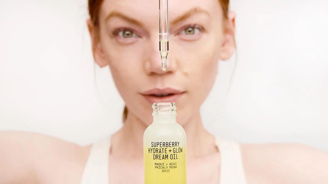 Close-up studio shot of a woman with red hair and pale skin presenting a bottle of Superberry Hydrate + Glow Dream Oil.