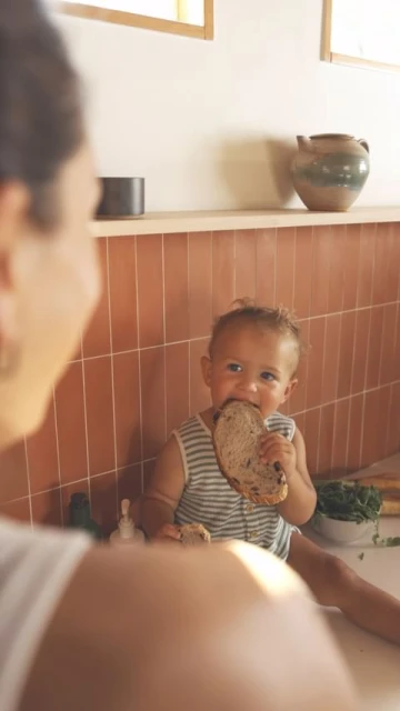 A baby sits on a kitchen counter eating a large slice of bread, with a woman in the foreground and a tiled backsplash behind.