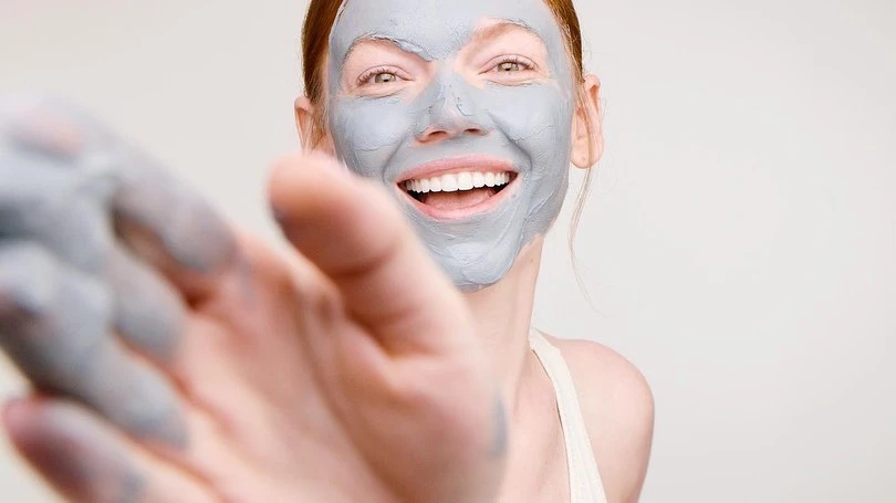 Smiling woman with grey face mask looks at the camera, holding out her hand with the same mask material on it, indoors.