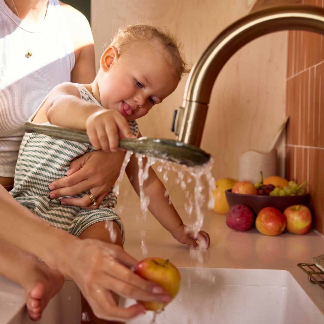 Child with adult playing with water from a kitchen faucet with fruit on counter. Kid is holding green vegetable under water.