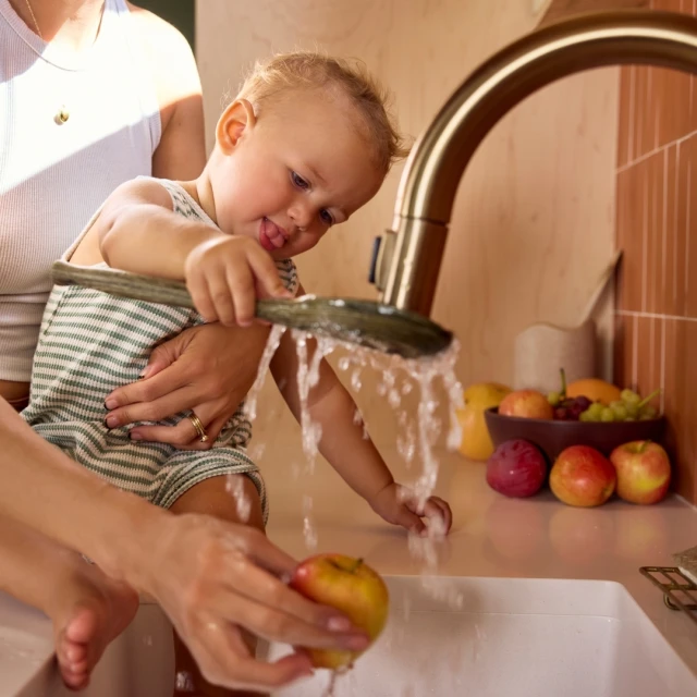Child with adult playing with water from a kitchen faucet with fruit on counter. Kid is holding green vegetable under water.