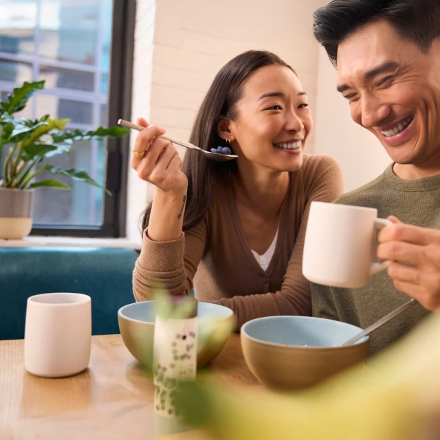 A smiling Asian couple at a table shares a morning meal. The woman holds a spoonful of blueberries, and the man holds a mug.