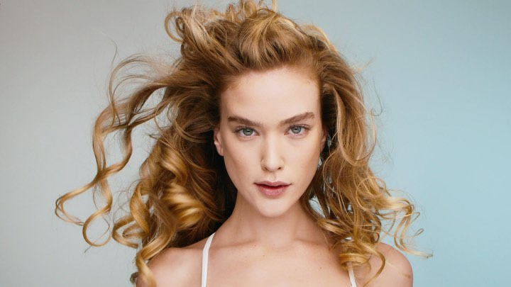 Close-up portrait of a beautiful adult woman with curly strawberry-blonde hair posing in a studio with a blue and gray background.