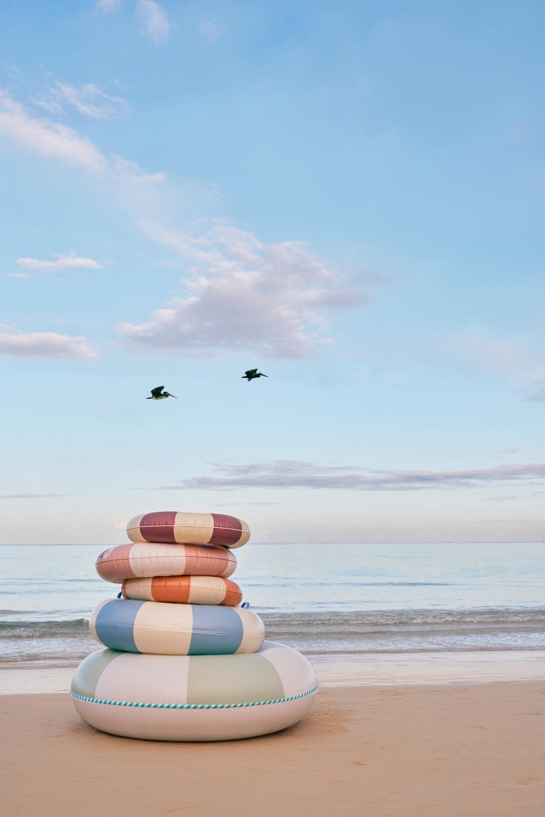 Stack of colourful striped inflatable swim rings on sandy beach with blue sea and sky backdrop, featuring birds flying above the water.
