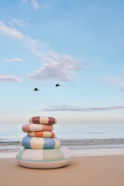 Stack of colourful striped inflatable swim rings on sandy beach with blue sea and sky backdrop, featuring birds flying above the water.