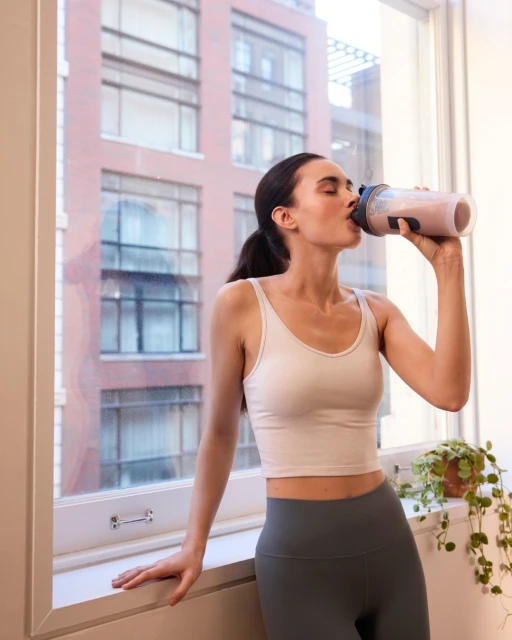 Adult woman in exercise clothing stands indoors drinking from a large bottle by a window with an urban building view.