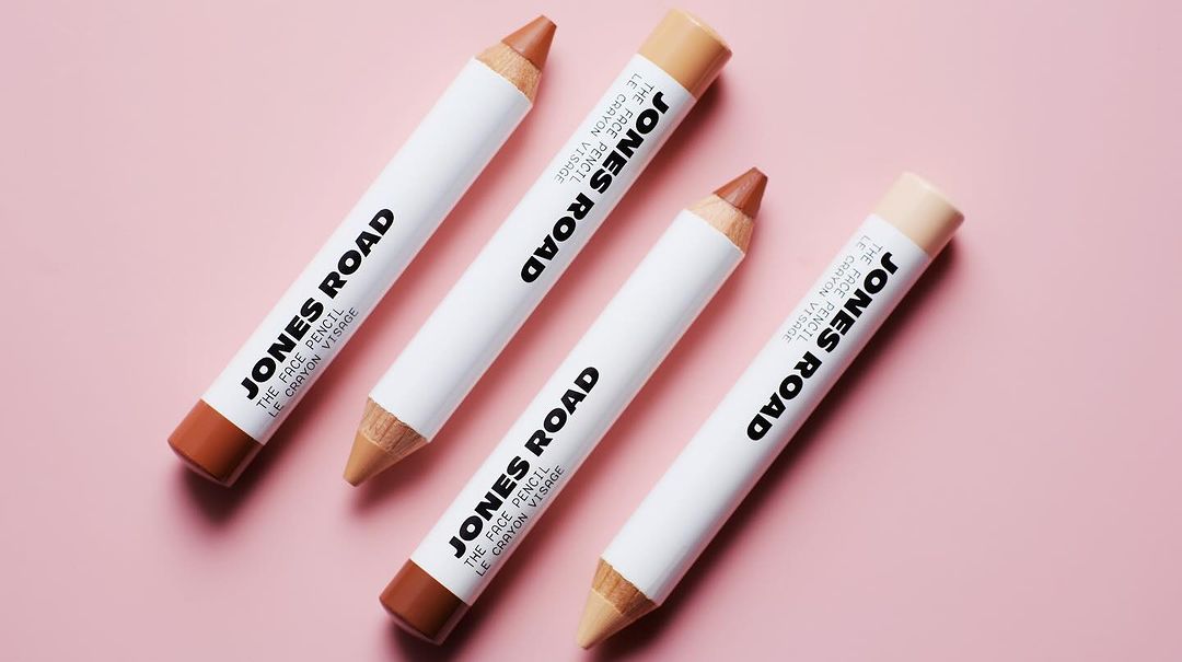 Four Jones Road face pencils in various neutral shades are arranged on a pink surface in a beauty product shot, showcasing their design.