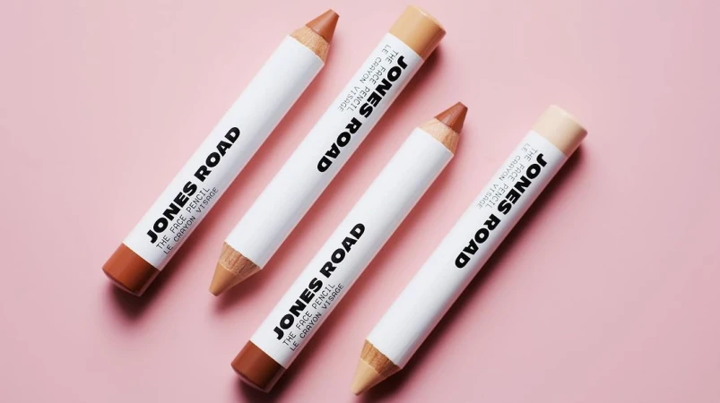 Four Jones Road face pencils in various neutral shades are arranged on a pink surface in a beauty product shot, showcasing their design.