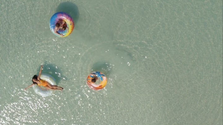Aerial view of three people floating in inflatable rings on calm, clear, light turquoise ocean water. Summer fun and relaxation.