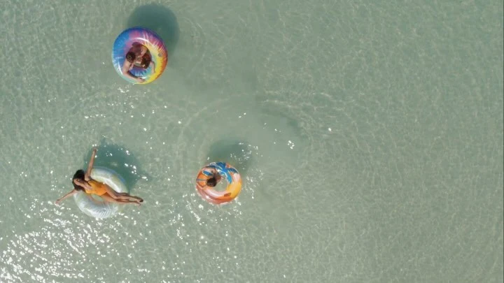 Aerial view of three people floating in inflatable rings on calm, clear, light turquoise ocean water. Summer fun and relaxation.