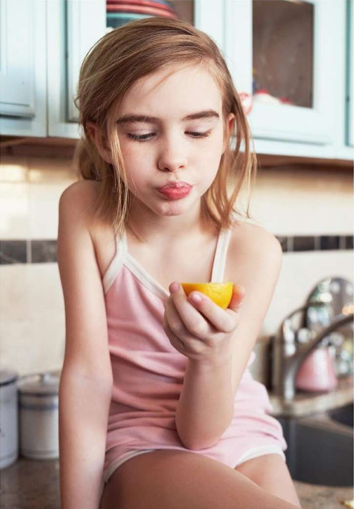 Young girl in a pink top holds a half lemon, puckering her lips in a kitchen setting with pale blue cabinets and neutral countertops.