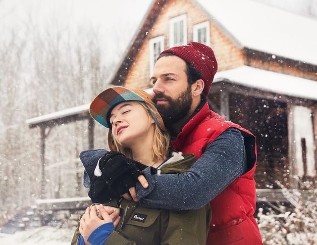 Couple in winter clothing embraces outside a rustic cabin during a snowfall, creating a warm and intimate scene in a cold environment.