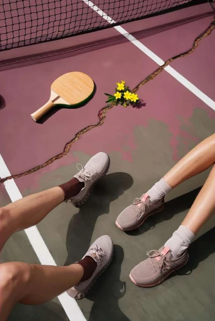 Overhead shot of a table tennis paddle, small yellow flowers, and two pairs of feet wearing athletic shoes on a purple and green court.
