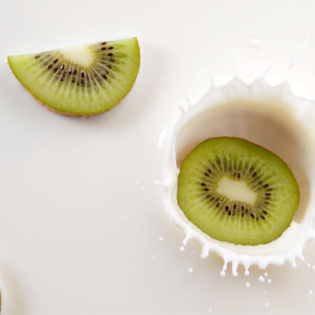Overhead studio shot of two kiwi fruit slices, one dropping and splashing into white milk, creating a dynamic, refreshing scene.