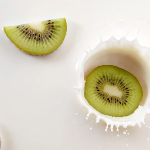 Overhead studio shot of two kiwi fruit slices, one dropping and splashing into white milk, creating a dynamic, refreshing scene.