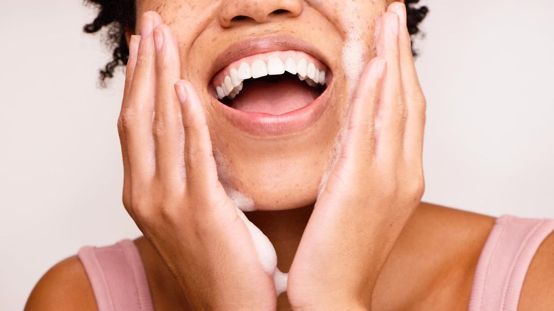 Close-up of a smiling, fair-skinned woman with dark curly hair washing her face with a foamy cleanser, set against a neutral backdrop.