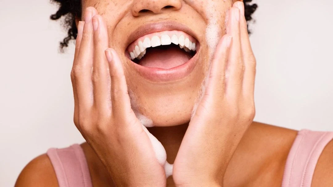 Close-up of a smiling, fair-skinned woman with dark curly hair washing her face with a foamy cleanser, set against a neutral backdrop.