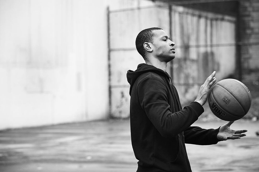 Black and white image of an adult male spinning a basketball on his finger, wearing a hoodie, in an outdoor urban environment.