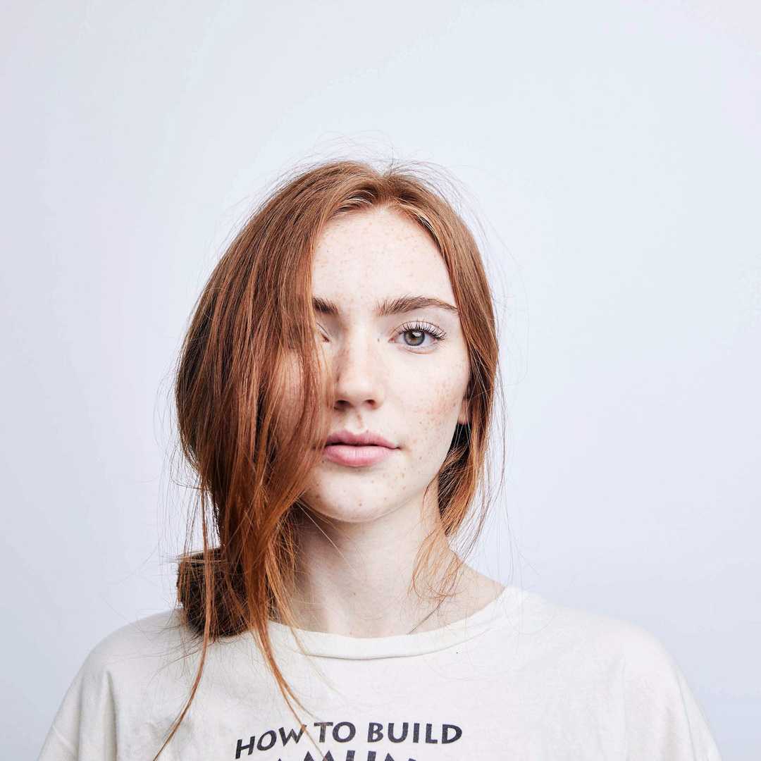 Close-up studio portrait of a young woman with freckles and red hair, wearing a graphic tee shirt, against a white backdrop.