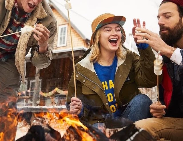 Three young adults laugh around a bonfire, toasting marshmallows on sticks near a rustic house in winter, creating a cozy outdoor scene.