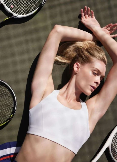 Adult woman lies on her back on a tennis court surrounded by tennis rackets, wearing a white crop top and blue striped skirt.
