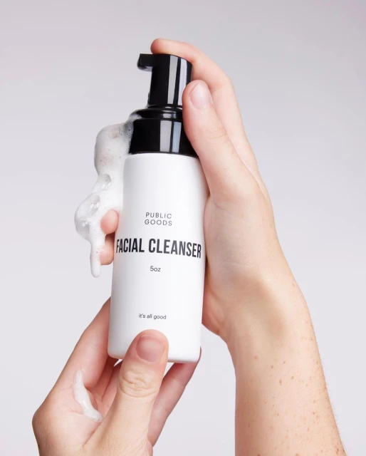Hands hold white bottle of foaming facial cleanser from Public Goods, black pump nozzle, product foams. Clean skincare product.