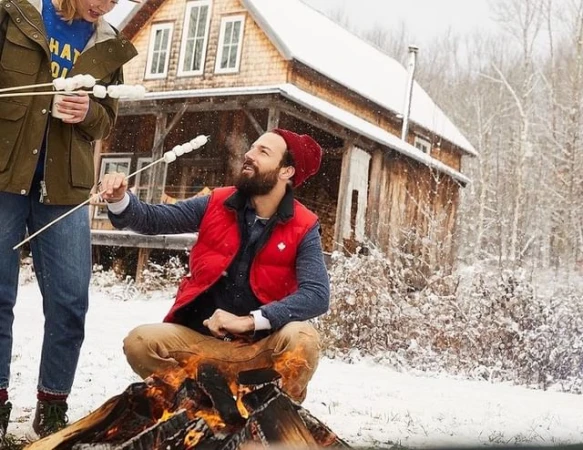 Man and woman roasting marshmallows over a campfire in a snowy landscape near a wooden cabin, wearing winter clothing and red beanie.