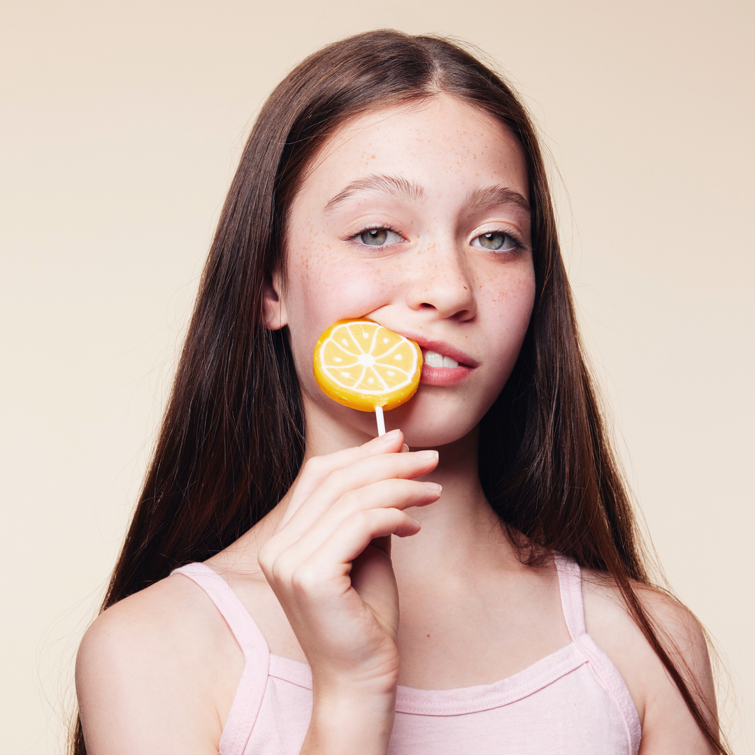 A young girl with long brown hair playfully bites a yellow lemon lollipop, set against a soft, neutral background in a vibrant studio shot.