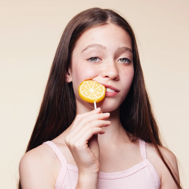 A young girl with long brown hair playfully bites a yellow lemon lollipop, set against a soft, neutral background in a vibrant studio shot.