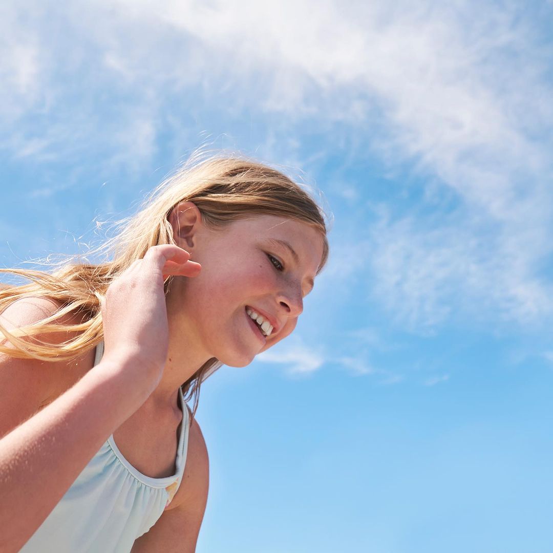 Young girl with blond hair smiles broadly against a bright blue sky with white clouds in an outdoor setting on a sunny day.