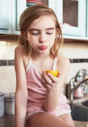Young girl in a pink tank top holding a lemon slice with a puckered expression in a kitchen