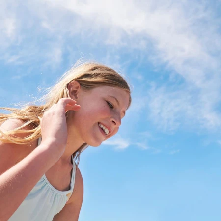 Young blond girl smiling against a bright blue sky with wispy clouds, shot from a low angle by Candy Kennedy.