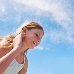 Young blond girl smiling against a bright blue sky with wispy clouds, shot from a low angle by Candy Kennedy.