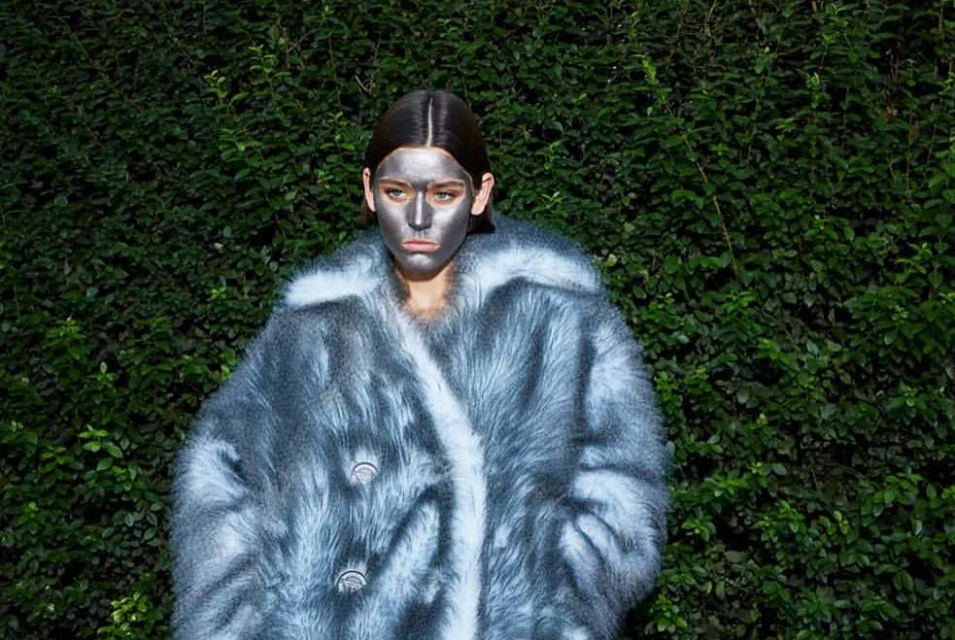 Woman with silver face paint wears furry blue and white coat, posing outdoors against a green leafy wall. Fashion and beauty shot.