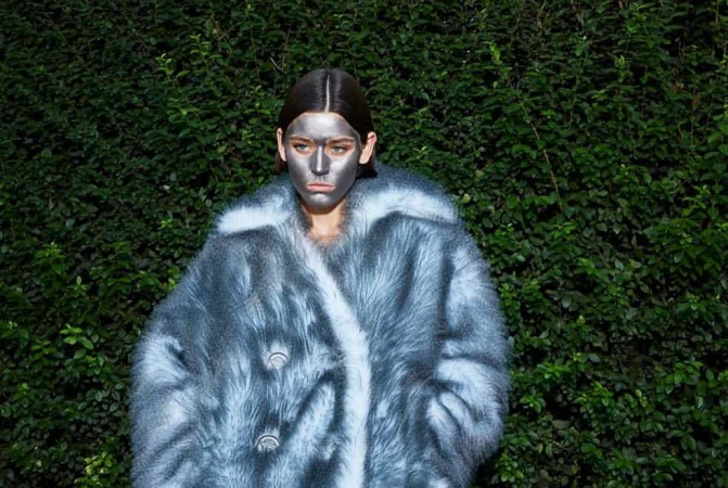 Woman with silver face paint wears furry blue and white coat, posing outdoors against a green leafy wall. Fashion and beauty shot.