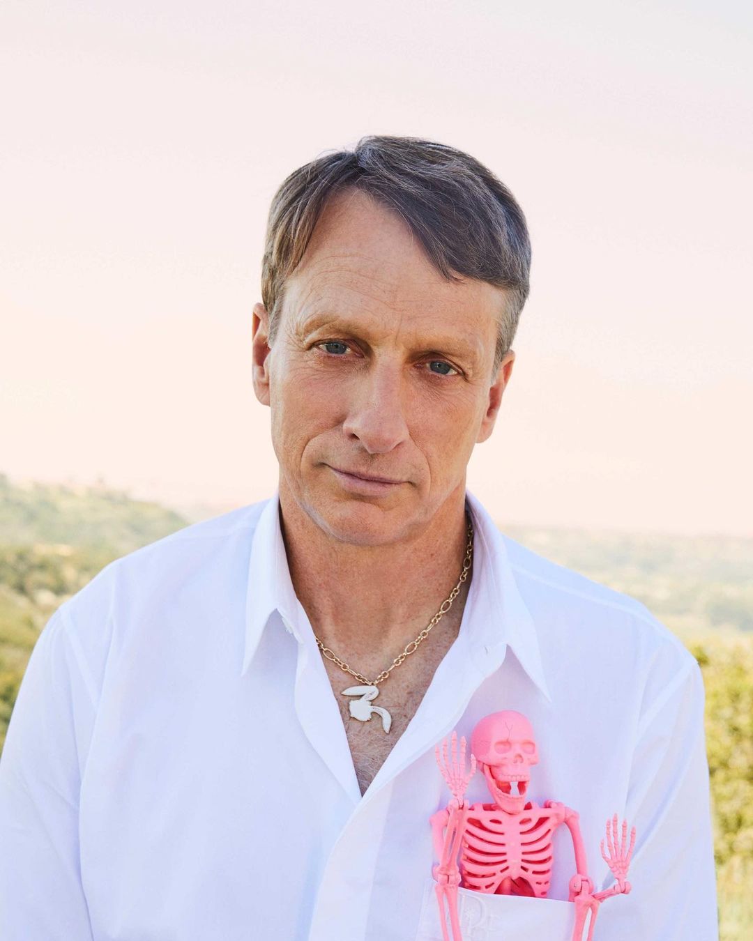 Portrait of Tony Hawk in a white shirt with a skeleton toy in his pocket. Soft, diffused light and a blurred, natural background.