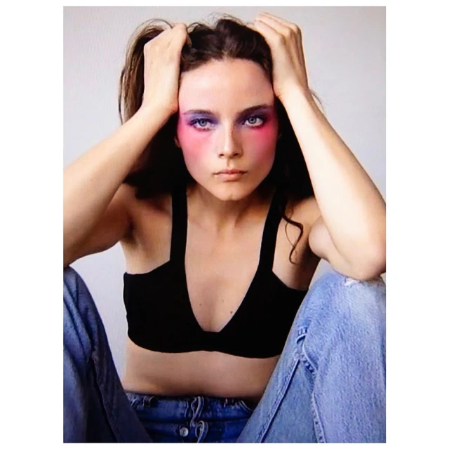 Eye-level portrait of a young woman with pink and blue eye makeup wearing a black top and jeans posing in front of a grey background.