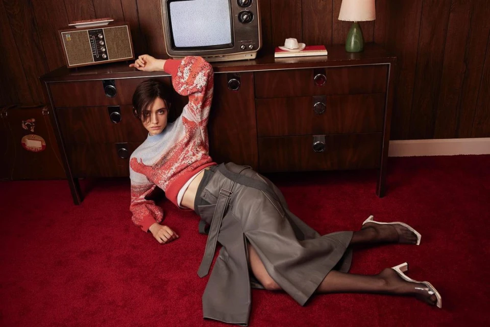 A woman poses on a red carpet wearing a sweater and skirt next to a wooden dresser with a vintage television and radio on top.
