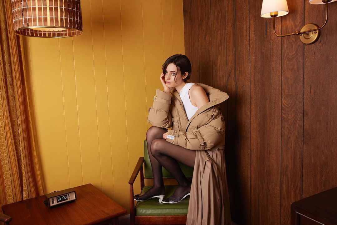 A woman with short dark hair sits on a green chair wearing a tan puffer jacket and black tights in a retro room with yellow walls.