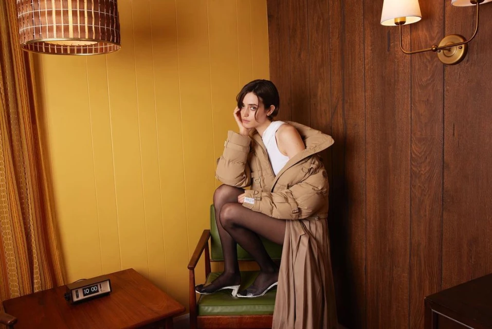 A woman with short dark hair sits on a green chair wearing a tan puffer jacket and black tights in a retro room with yellow walls.