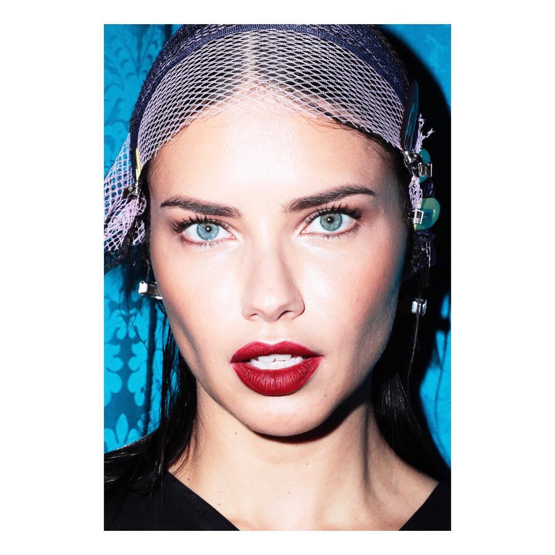 Close-up studio portrait of a woman with red lipstick and blue eyes wearing a net head covering against a blue patterned background.