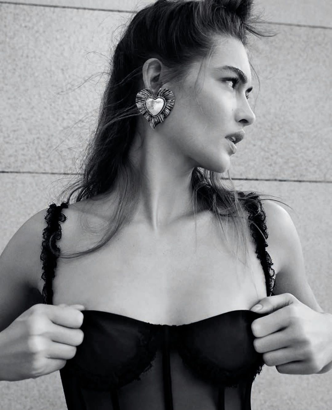 Black-and-white portrait of a young adult woman with styled hair, heart-shaped earrings, and a black lace top against a textured wall.