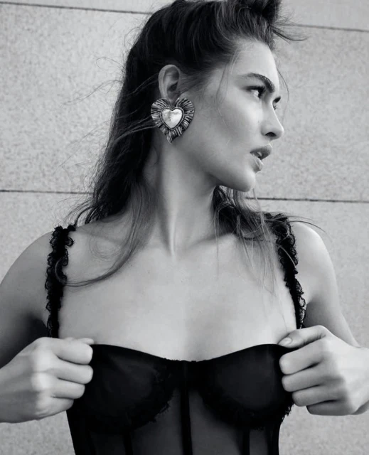 Black-and-white portrait of a young adult woman with styled hair, heart-shaped earrings, and a black lace top against a textured wall.