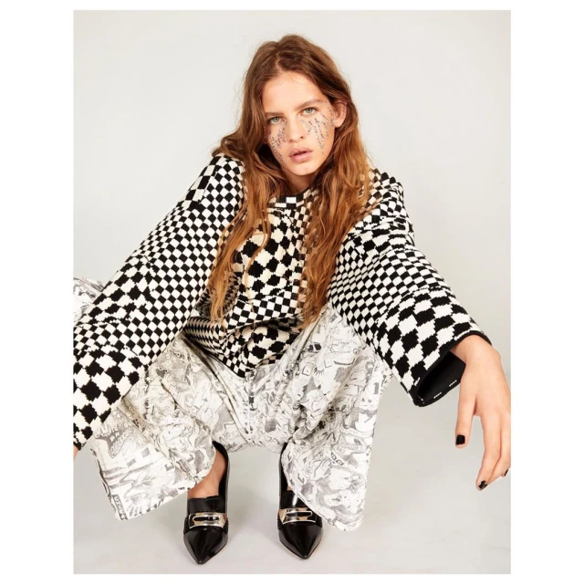 A young adult woman in a black-and-white checkered shirt and patterned pants poses in a studio with dark shoes and glitter on her face.
