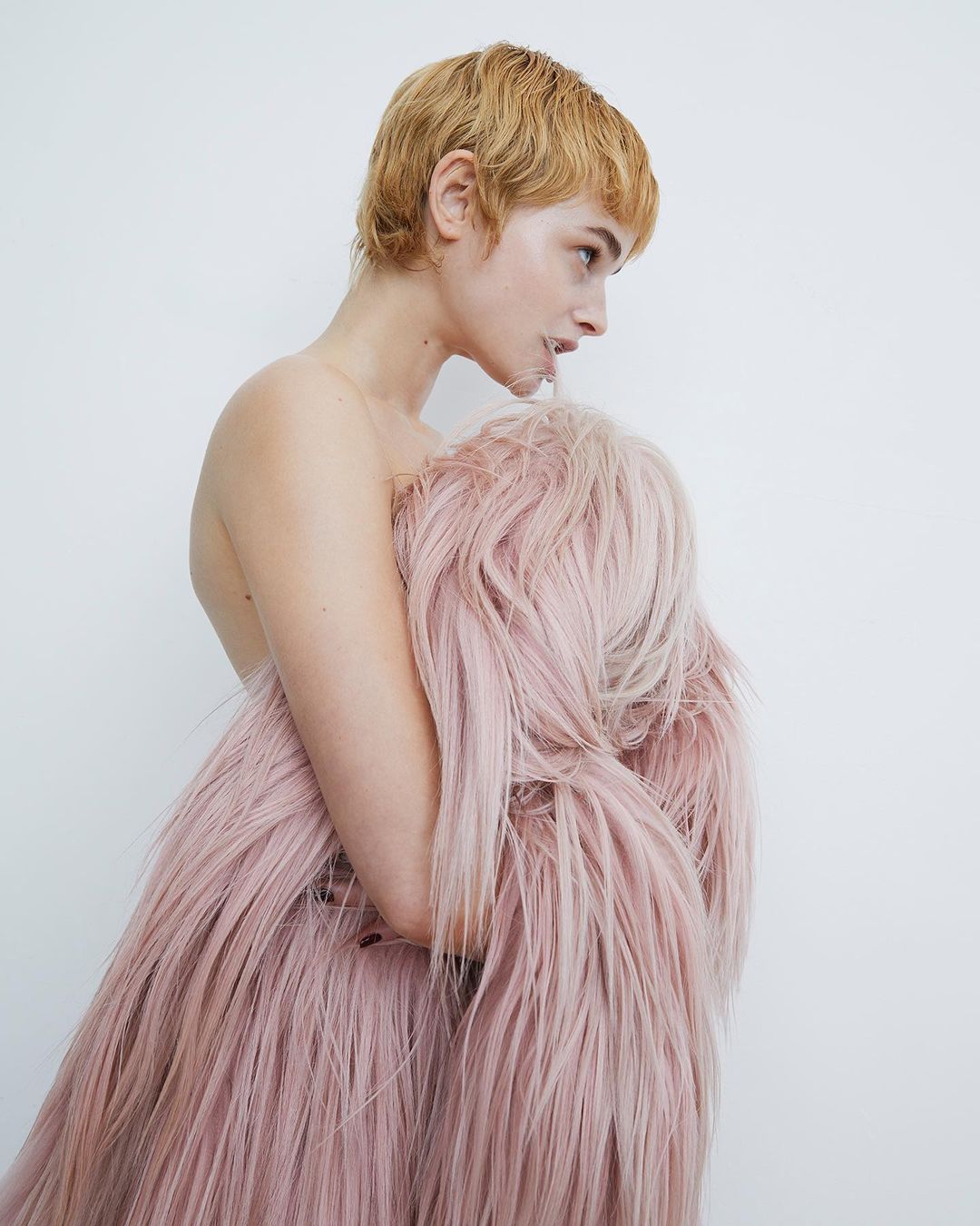 Woman with blonde pixie cut poses sideways partially covered by a long pink faux fur coat in a brightly lit photo studio.