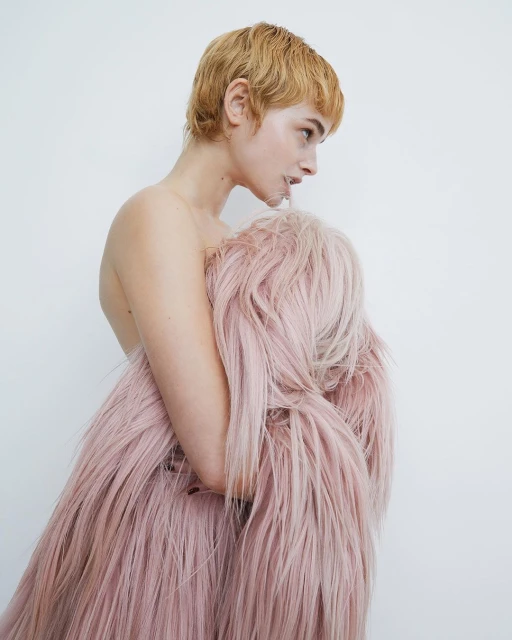 Woman with blonde pixie cut poses sideways partially covered by a long pink faux fur coat in a brightly lit photo studio.