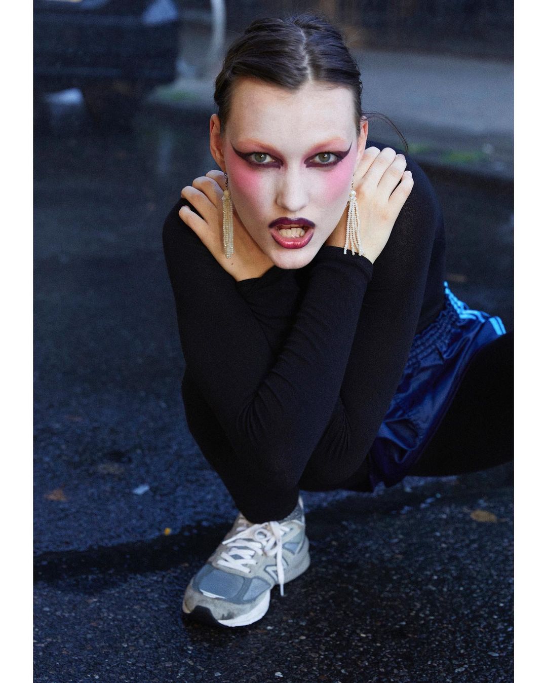Young woman crouches on a wet urban street in black top, blue shorts, gray sneakers, pink eye shadow, and long earrings.